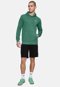 Felpa verde con tasca frontale, cordini e logo. Abbinata a pantaloni corti neri e sneakers verde menta con dettagli bianchi e logo.