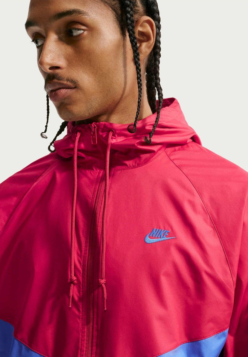 Man met gevlochten haar die een felrode Nike-jas met blauw logo draagt en opzij kijkt tegen een effen lichte achtergrond.