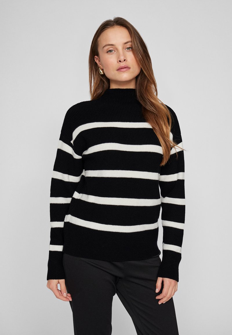 Pulover cu guler înalt, dungi alb-negru, cu textură ribbită, mâneci lungi și o croială lejeră, asortat cu pantaloni negri.