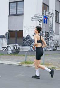 Un soutien-gorge de sport noir et un short assorti, portés avec des chaussures de course noires et des chaussettes blanches, par une personne qui jogge dans une rue de la ville.