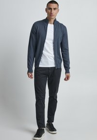 Marine glidelås kardigan med ståkrage, laget av et lett blandingsstoff, brukt over en hvit t-skjorte og kombinert med mørke bukser og svarte joggesko.
