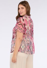 Blusa transparente con un colorido patrón ikat en rosa, turquesa y beige, con mangas cortas con volantes y un corte relajado.