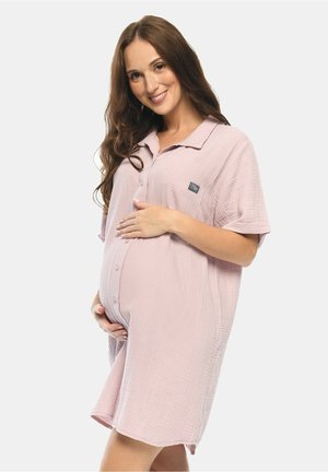 Schwangere Frau lächelnd, trägt ein lockeres, hellrosa Hemdkleid, hält ihren Bauch sanft mit beiden Händen vor einem einfarbigen Hintergrund.