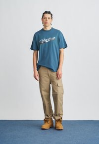 Camiseta azul de manga corta con logo metálico, pantalones cargo color beige y botas marrones, de pie contra un fondo liso.