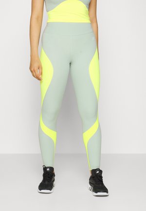 Leggings de sport vert clair avec des panneaux latéraux jaune fluo, taille sans couture et design ajusté. Portés avec des chaussures de sport noires.