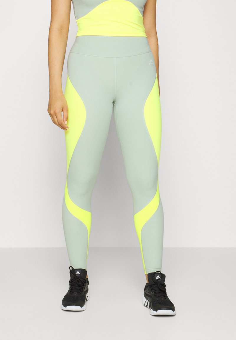 Leggings sportivi verde chiaro con pannelli laterali giallo neon, vita senza cuciture e design aderente. Indossati con scarpe sportive nere.