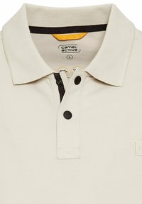 Beige bomulds polo skjorte med ribbet krave, to sorte knapper og et broderet logo på brystet. Har kontrasterende mørke detaljer.