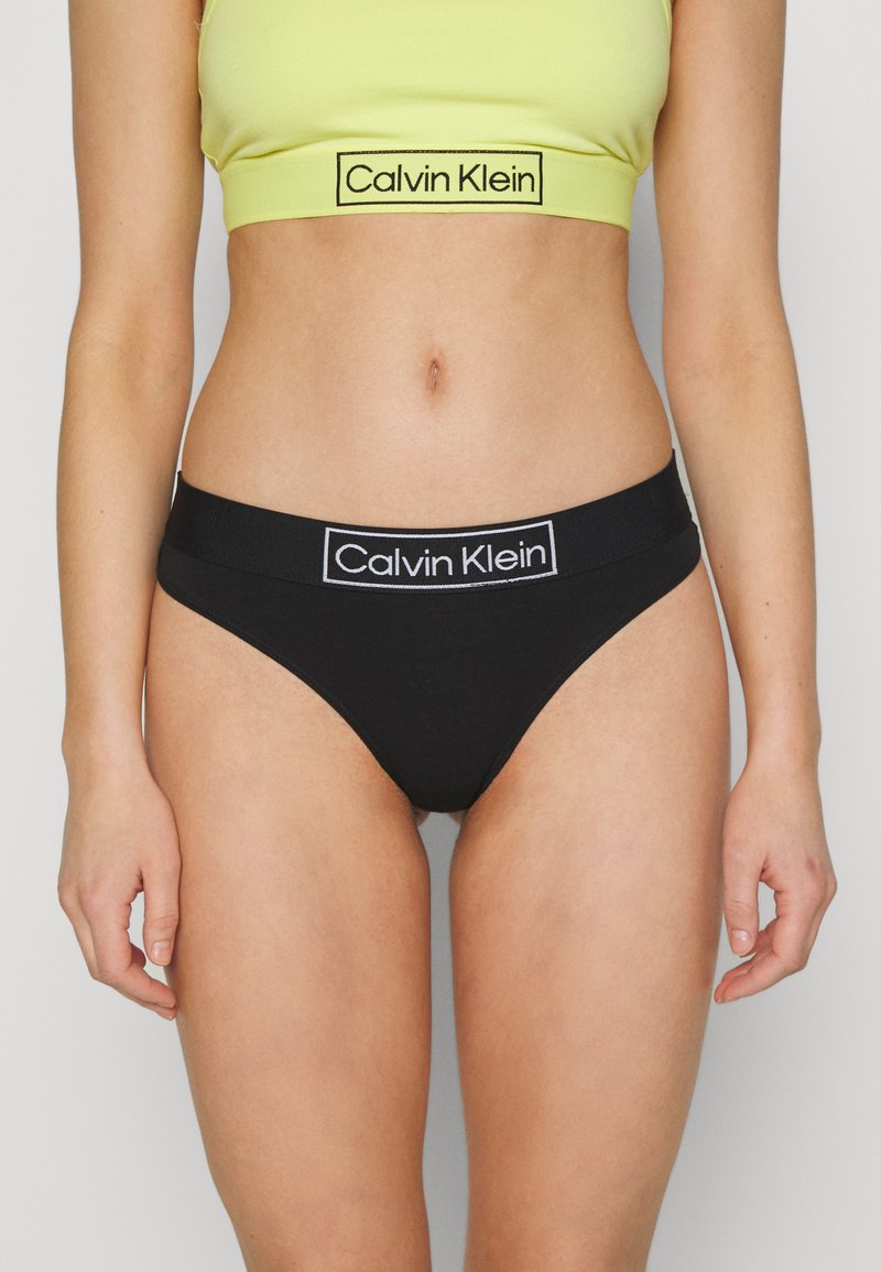 Culotte bikini en coton noir avec une large ceinture élastique arborant un logo "Calvin Klein" en blanc. Le haut est un débardeur cropped ajusté jaune.