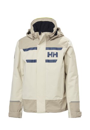 Niño con chaqueta impermeable beige y marrón claro con capucha y pantalones cortos azul marino, mirando hacia un lado contra un fondo liso.