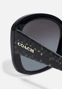 Ochelari de soare negri cu o finisare lucioasă, formă pătrată supradimensionată și lentile cu un gradient. Au un braț texturat cu marcajul „COACH” de culoare aurie.