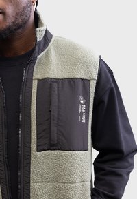 Man in een lichtgrijze fleece vest met een donkergrijze ritszak op de borst, gedragen over een zwart langemouwen shirt, waarbij de linkerkant van de torso en de rand van het gezicht zichtbaar zijn.