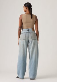 Γυναίκα με πλεξούδες, φορώντας μπεζ αμάνικη μπλούζα και ανοιχτό μπλε τζιν παντελόνι με φαρδιά πόδια, στέκεται γυρισμένη με την πλάτη σε απλό φόντο.