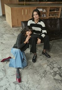 Deux femmes sont assises sur un canapé en cuir brun foncé. L'une porte un haut rayé noir et blanc et un pantalon noir ; l'autre porte un haut gris, un jean bleu et des chaussures rouges.