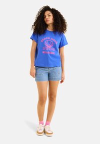 Camiseta de algodón azul con un gráfico rosa que dice "PLAYA EN HUMOR"., combinada con pantalones cortos de mezclilla y calcetines rosa claro. Estilo de zapatillas estándar.