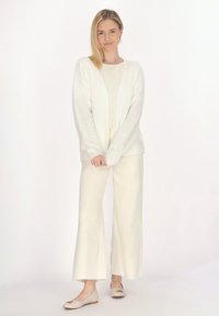 Witte fluffy cardigan, crèmekleurig top en wijdvallende crèmekleurige broek. Eenvoudige ronde beigekleurige ballerina's met een kleine decoratieve accent. Neutraal kleurenpalet.