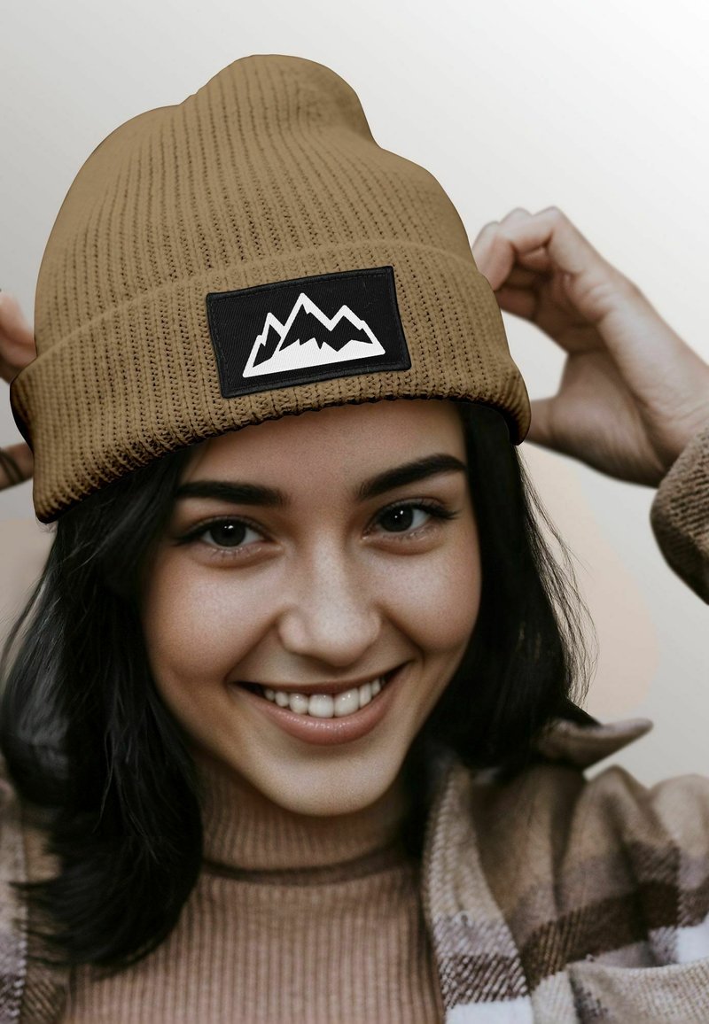 Brown ribbed knit beanie with a black patch featuring white mountain graphics. The texture is soft, and the design is form-fitting.