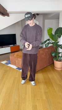 Maglione marrone lavorato a maglia spesso con scollo ampio, abbinato a pantaloni di velluto a coste scuri. Scarpe da ginnastica argento e un berretto nero completano l'outfit.