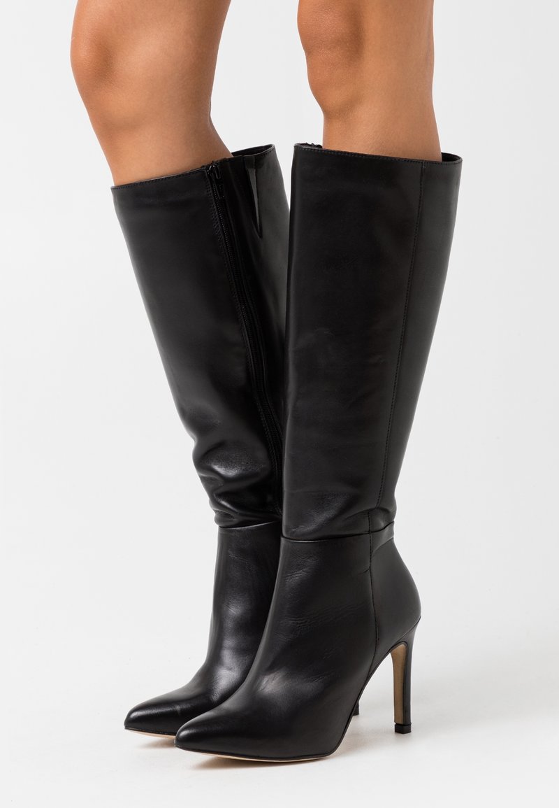 Bottes noires en cuir montantes jusqu'aux genoux avec talons et fermetures éclair latérales, portées sur des jambes nues, présentées sur un fond clair uni.