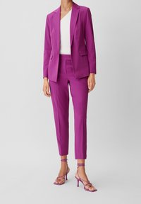 Costume fuchsia avec un blazer ajusté et un pantalon cintré, associé à des talons hauts à brides. Le tissu semble lisse, avec un design moderne et élégant.