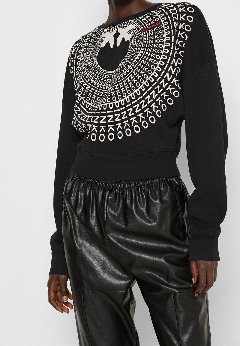 Černá krátká mikina s bílým geometrickým grafickým motivem a nápisy. Kombinováno s lesklými černými plisovanými kalhotami.