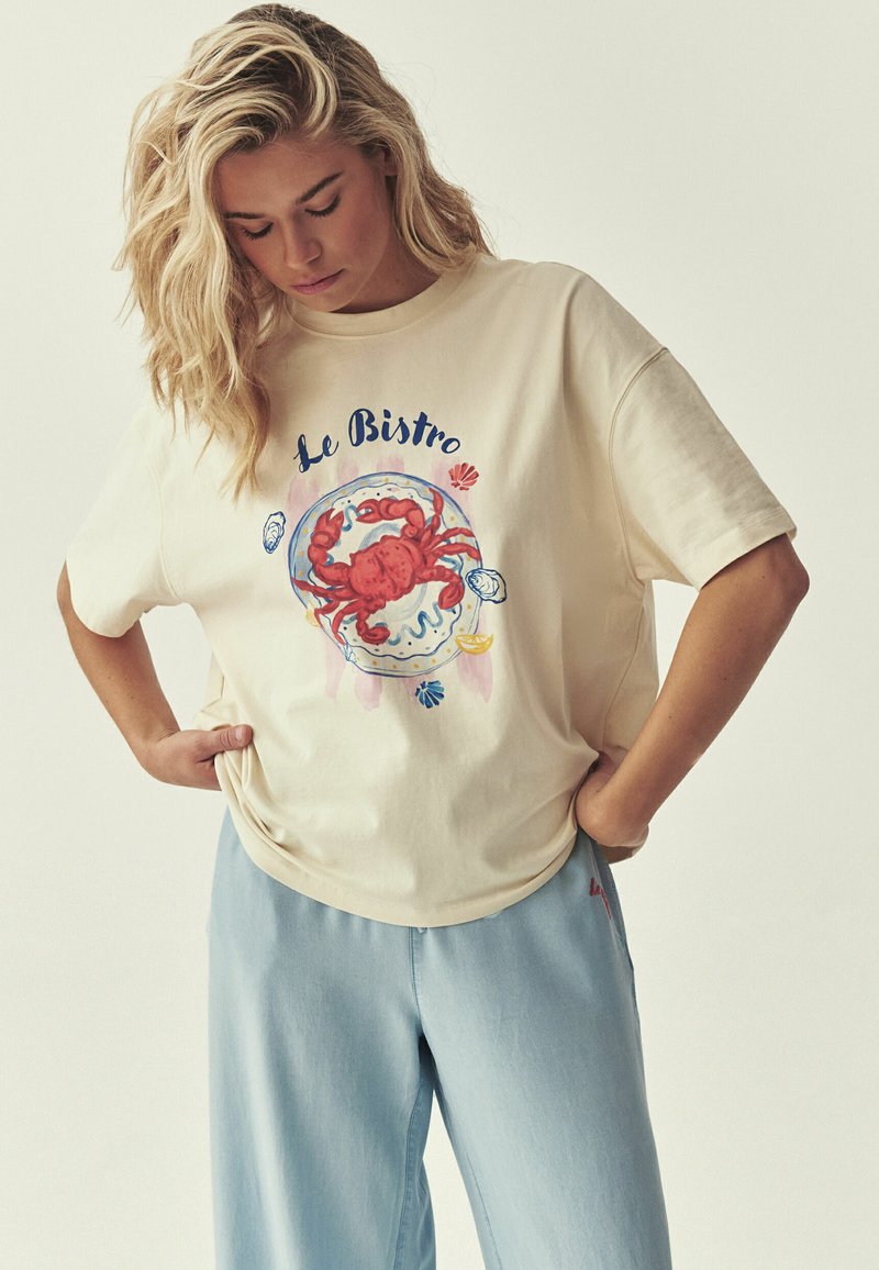 Femme blonde regardant vers le bas, portant un t-shirt crème avec un crabe rouge et le texte « Le Bistro », associé à un pantalon bleu clair, mains dans les poches.