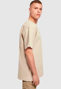 Beige bavlněné tričko s krátkým rukávem, volný střih, kulatý výstřih a rovný spodní lem. Nosené s světlými kalhotami, zobrazené z boku.