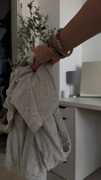 Des vêtements gris côtelés sont tenus dans une main, montrant une texture douce et un design ample. Une main avec des bracelets en perles est visible au premier plan.