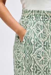 Hand in pocket wearing green and white patterned pants with elastic waistband and white sleeveless top.