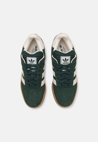 Green suede sneakers with white stripes and a brown rubber sole. Lace-up design, cushioned collar, and textured tongue with branding.