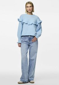 Svetlo modra pletena pulover s volančki na robu in rokavih, v kombinaciji z visokim pasom svetlim traper hlačami in športnimi supergami.