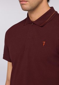 Poloko s krátkým rukávem v hnědé barvě vyrobené z texturované látky, s dvouknoflíkovou légou a oranžovým vyšitým akcentem na límci.