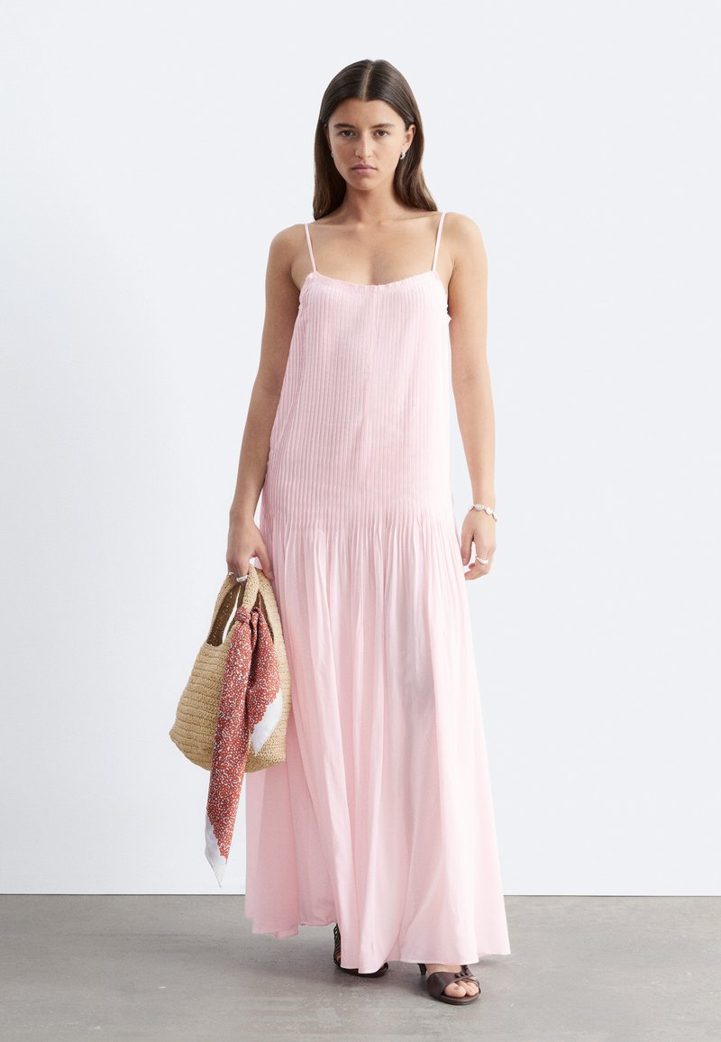 Femme aux longs cheveux foncés porte une robe maxi plissée rose clair et tient un sac cabas en paille avec un foulard à motifs.