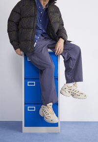 Giacca puffer con un motivo paisley scuro, camicia blu con bottoni, pantaloni grigi e sneakers beige con design a ritaglio, posizionati su un mobile blu.