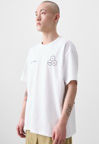 White short-sleeve t-shirt featuring hexagonal graphic with "ALL CONDITIONS GEAR." Smooth cotton fabric, relaxed fit, and no notable patterns.