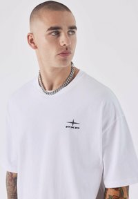 Camiseta de algodón blanca con cuello redondo y mangas cortas, que presenta un logotipo negro en el lado izquierdo del pecho. Incluye un collar de cadena plateada.