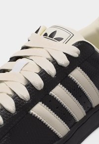 Ασπρόμαυρο sneaker Adidas με υφή μαύρου δέρματος, τρεις λευκές ρίγες, λευκά κορδόνια και λογότυπο Adidas στη γλώσσα.