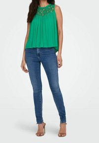 Blusa verde senza maniche con pieghe e dettagli in pizzo sul collo, abbinata a jeans skinny blu e sandali con tacco trasparente.
