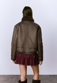 Veste en cuir marron avec un col en fausse fourrure prominent, cintrée à la taille, vue de dos, associée à une jupe bourgogne à plusieurs couches.