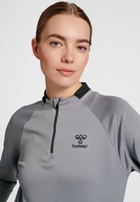 Maglietta grigia chiara a maniche lunghe con colletto a zip, caratterizzata da un logo nero e dettagli a contrasto. Il tessuto ha una finitura strutturata, adatta per l'abbigliamento sportivo.