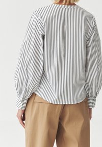 Valkoinen paita, jossa on mustia pystysuoria raitoja, kuhmurat kädet ja napitettu hihansuu; yhdistettynä vaaleisiin korkeavyötäröisiin housuihin.