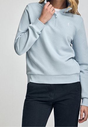 Femme portant un sweat-shirt bleu clair à fermeture éclair jusqu'au cou et un pantalon foncé, une main près de la fermeture éclair du col et l'autre le long du corps.