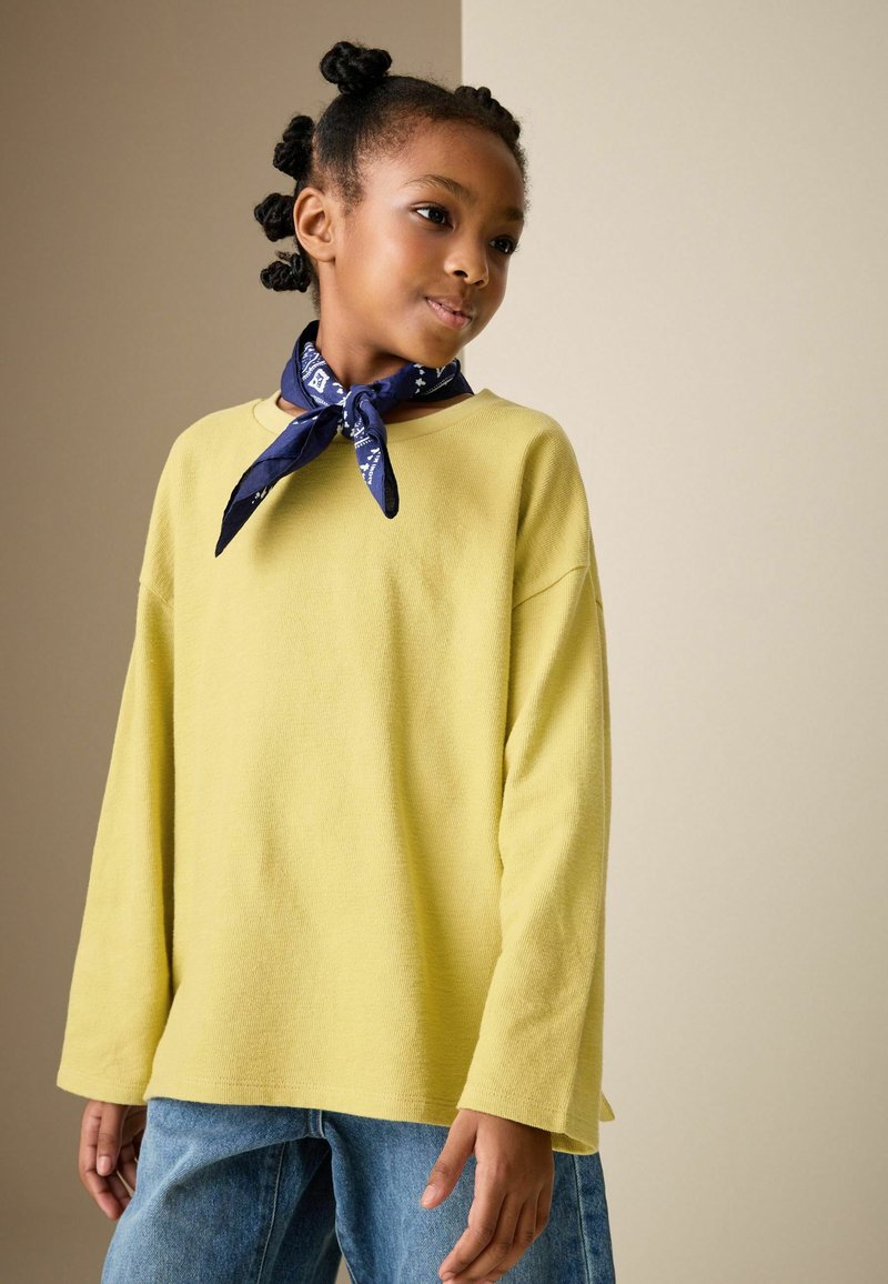 Top giallo a maniche lunghe con vestibilità rilassata, tessuto a coste e scollatura rotonda, abbinato a jeans di denim blu e a una sciarpa blu navy con fantasia.