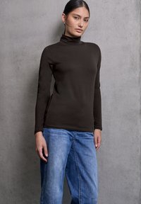 Jonge vrouw met donker haar in een knot, gekleed in een zwarte coltrui en losse blauwe jeans, staat voor een achtergrond van een grijze betonnen muur.