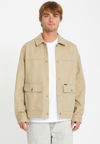 Chaqueta beige clara abotonada de tejido resistente, con cuello, dos bolsillos frontales y botones a presión plateados, llevada sobre una camisa blanca.