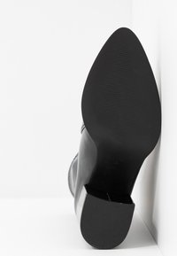 Svart høyhælte støvler med spiss tå, glatt materiale, teksturert såle og blank finish. Har firkantet hæl og kontrasterende sømmer.