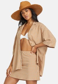 Checkered brown and cream outfit with a loose shirt and fitted skirt, complemented by a wide-brim straw hat.
