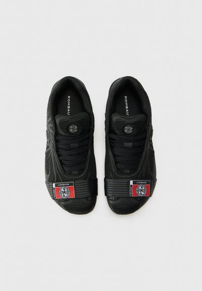 Baskets noires avec un dessus en mesh texturé, lacets plats, et une sangle proéminente arborant un patch logo rouge et noir sur le côté.