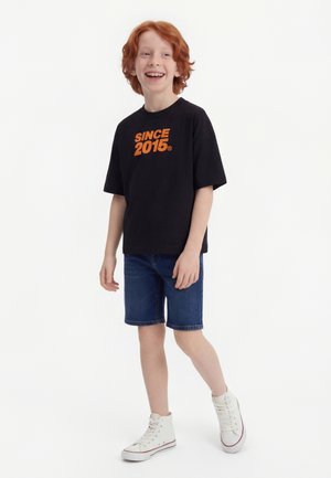 Garçon aux cheveux roux portant un t-shirt noir "SINCE 2015", un short en jean et des baskets blanches hautes, souriant et debout sur un fond blanc.