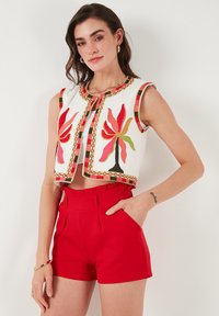 Gilet court blanc brodé avec un motif floral en rouge, rose et vert, associé à un short taille haute rouge avec poches latérales.