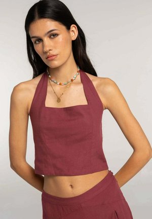 Joven mujer con cabello largo y oscuro que lleva un top corto burdeos sin mangas con cuello halter y falda a juego, complementada con collares en capas de cuentas y oro.